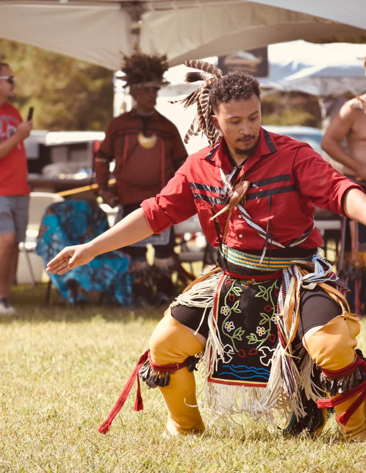 A man in a red ribbon shirt and beaded floral regalia dancing on the grass at the 2026 powwow